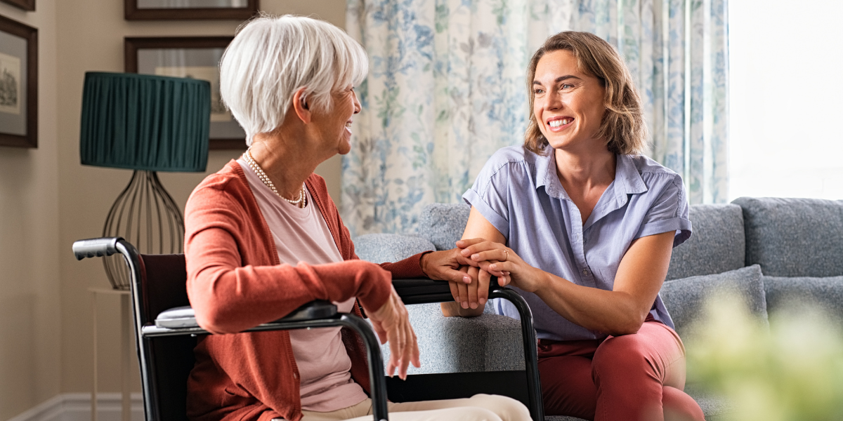 A woman, smiling and sitting on a couch, holds hands with her elderly mother in a wheelchair as they discuss assisted living.