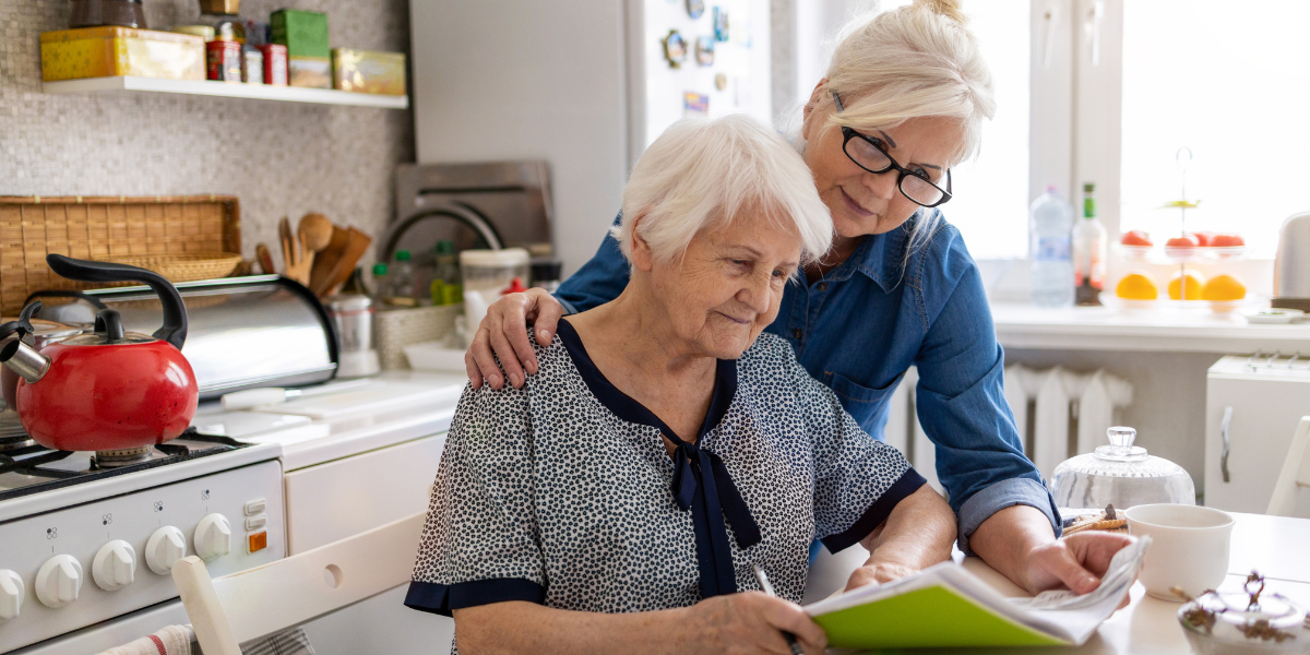 Daughter helping her elderly mother with assisted living paperwork at kitchen table.