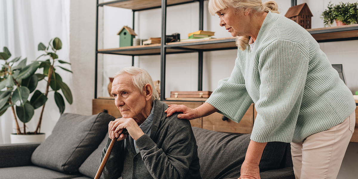 senior wife standing near retired husband sitting on sofa