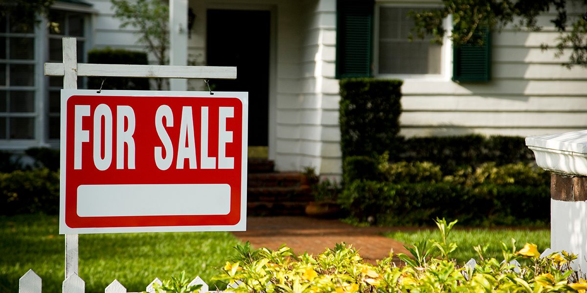 A red and white "For Sale" sign stands prominently in front of a well-maintained suburban home on a sunny day.